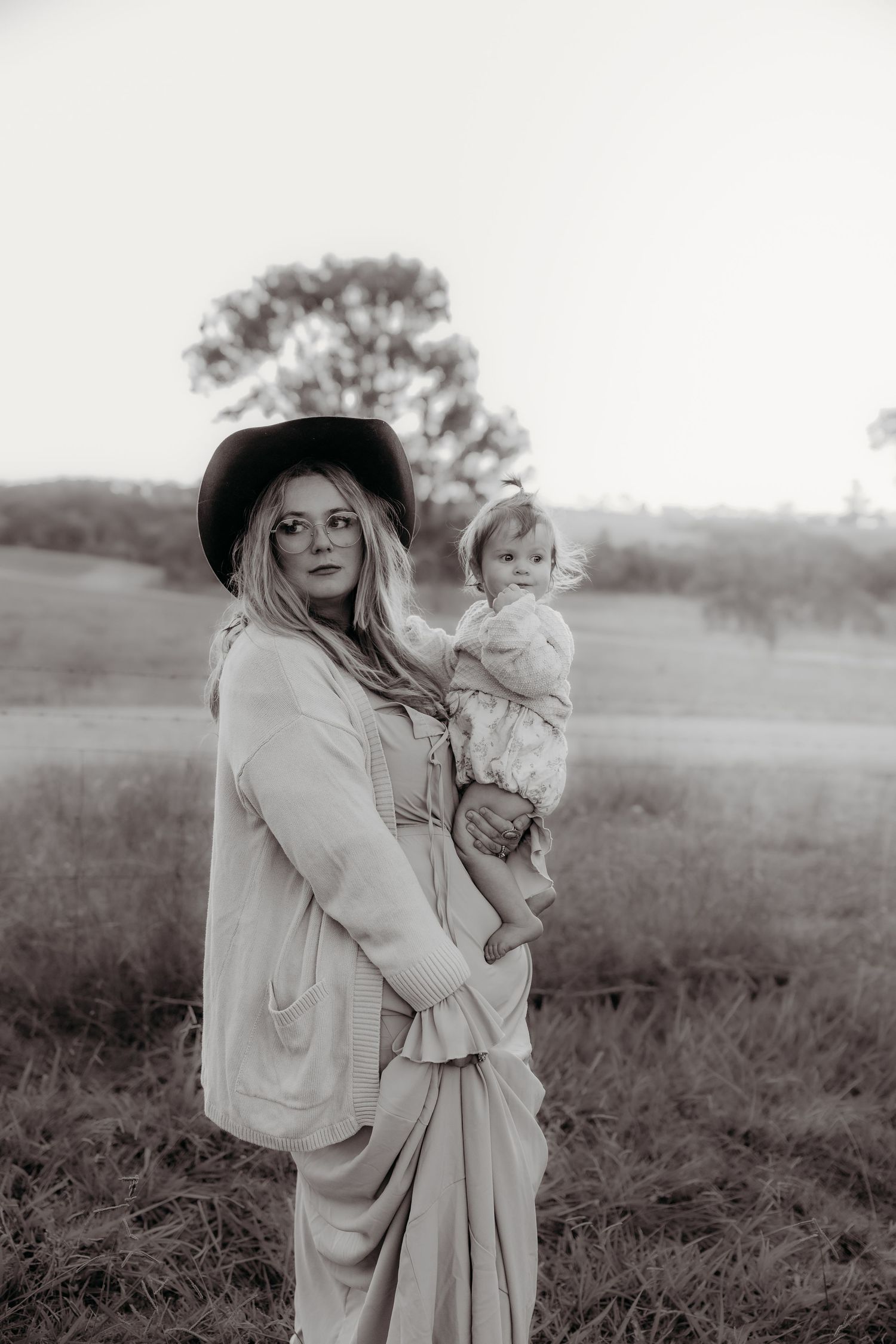 Black and white photo of a woman in a wide-brim hat holding a baby in a scenic rural field.