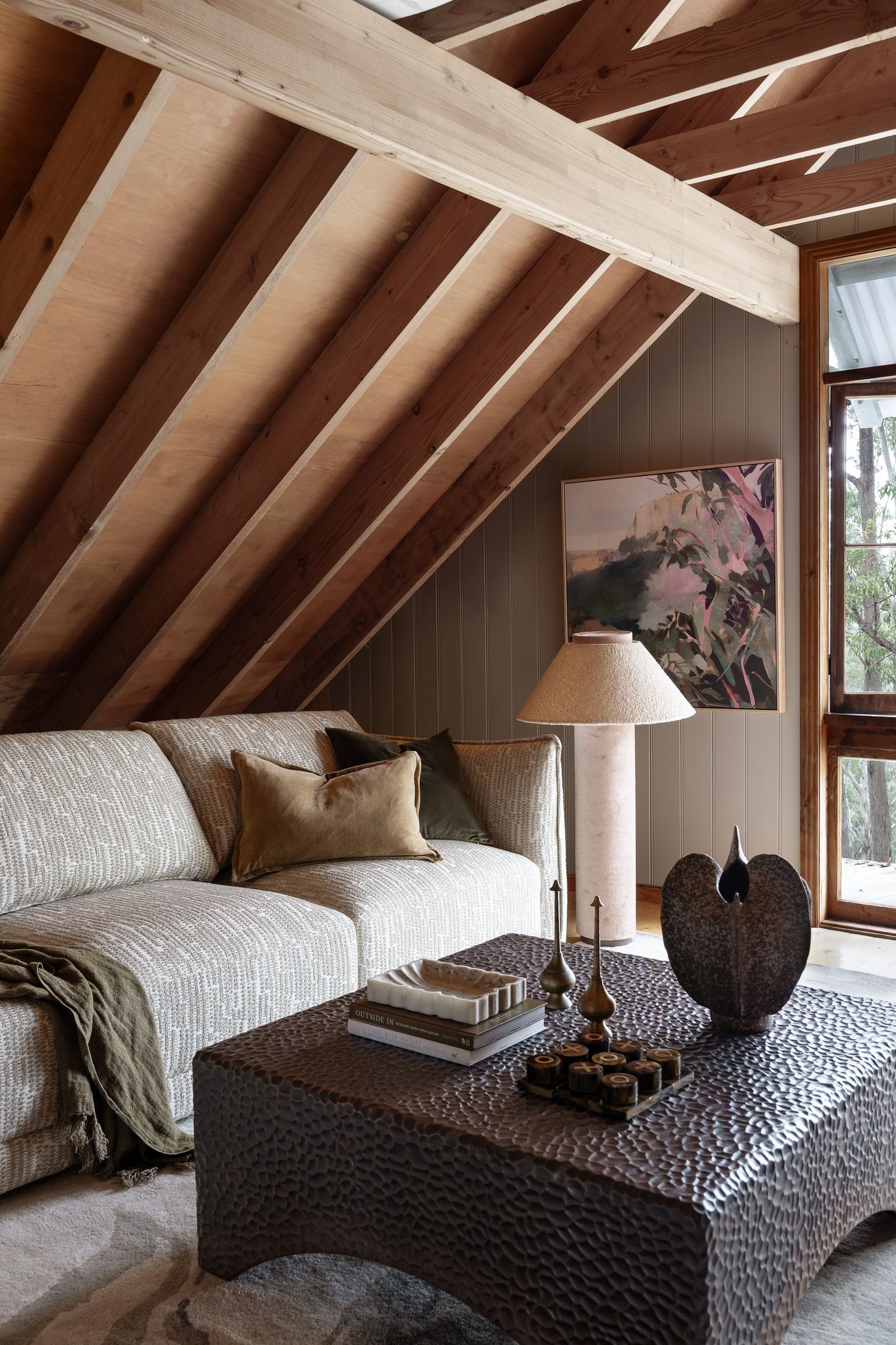 Cozy attic living room with exposed wood beams, cream sofa, textured dark coffee table, brass lamp, and floral artwork.