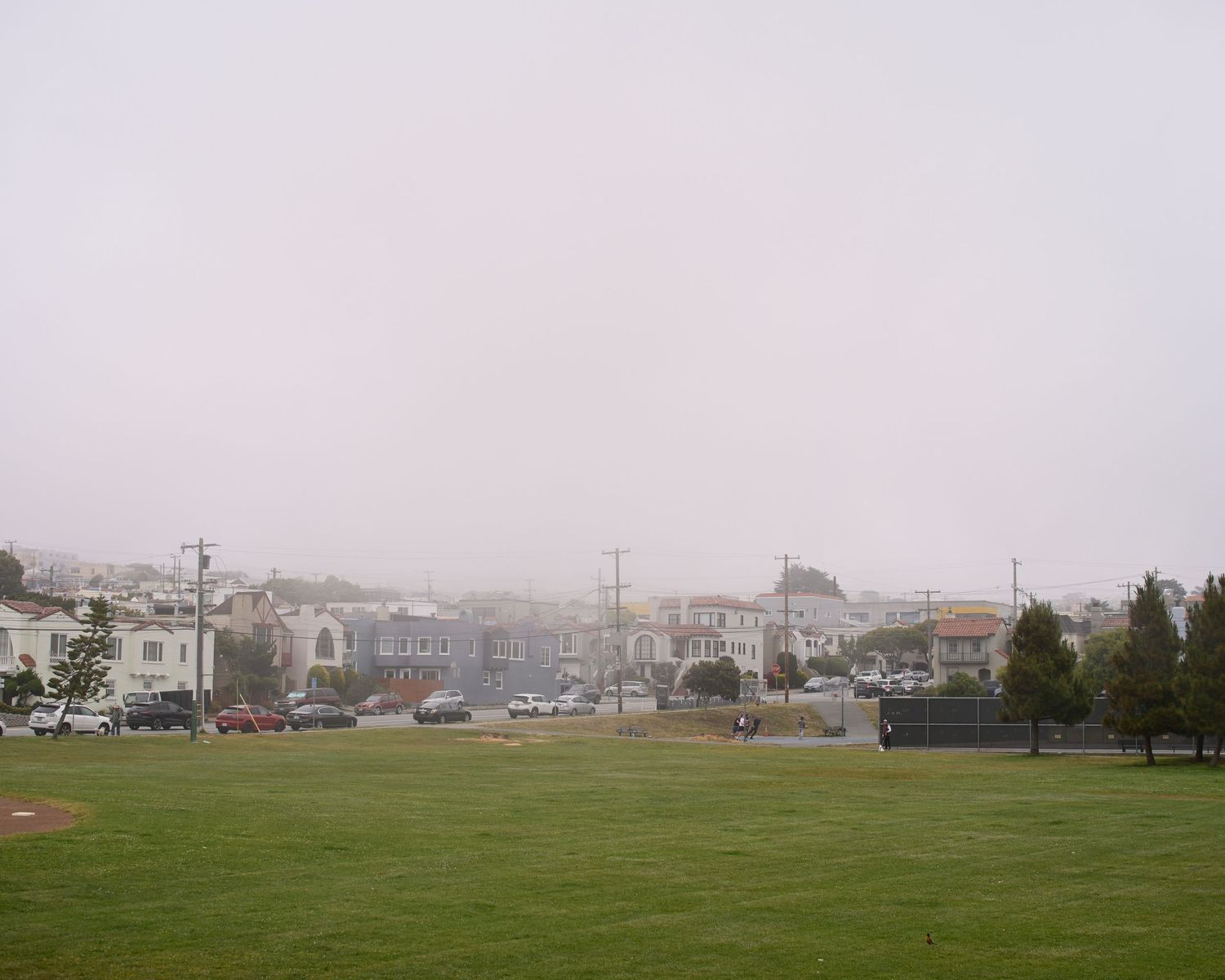 Foggy urban neighborhood with green grass field in foreground, residential buildings and utility poles visible in background.