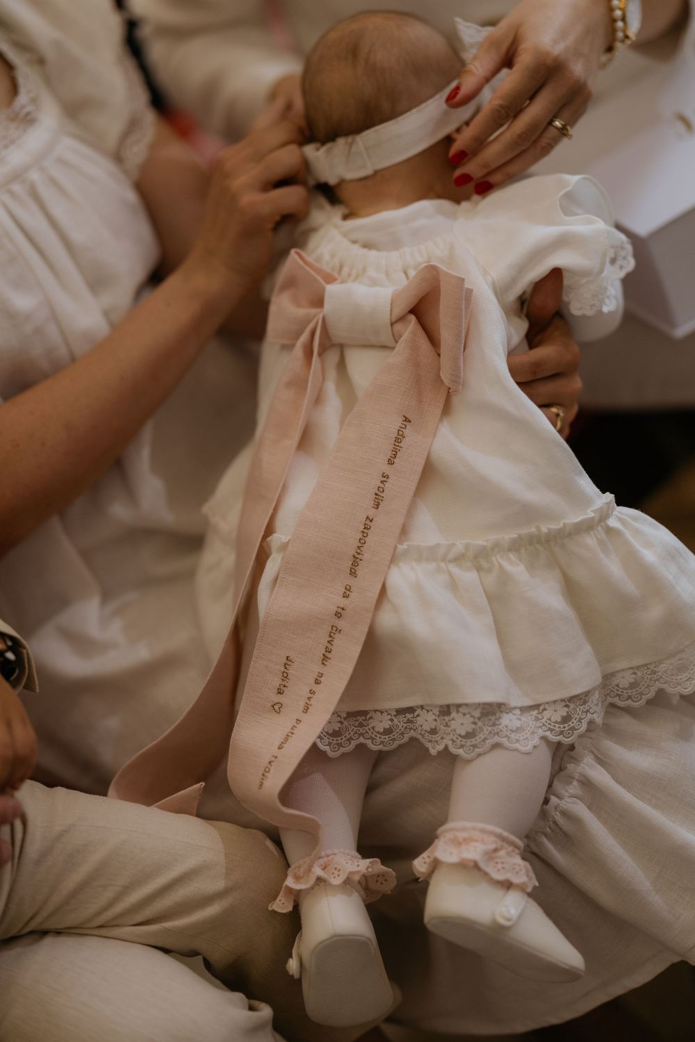 Baby in white lace christening dress with pink satin ribbon bow being dressed by family members.