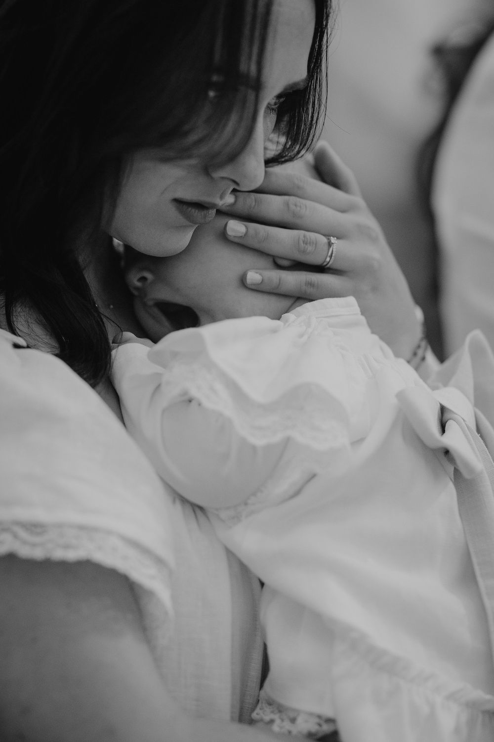 Tender black and white photo of a mother lovingly kissing her baby wearing a white christening gown.