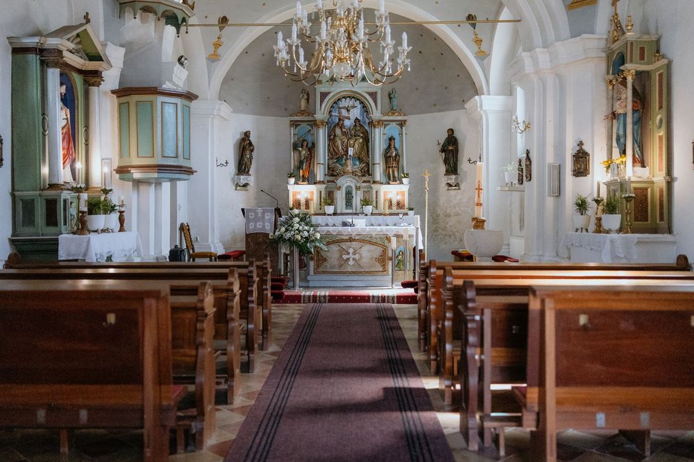 Elegant white Catholic church interior with wooden pews, ornate altar, chandelier, and red carpet aisle.