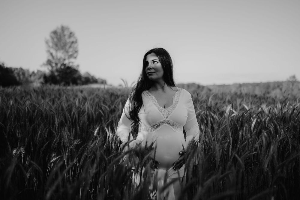 Black and white photo of a pregnant woman in a white dress standing in a tall grass field, cradling her belly.