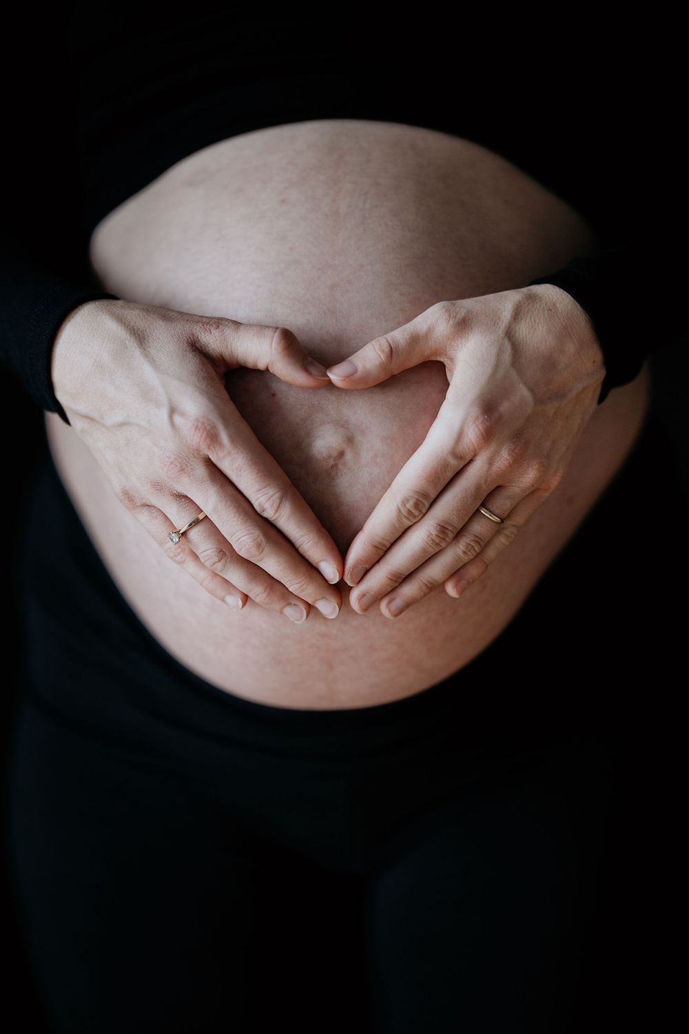 Pregnant woman forming a heart shape with her hands on her bare baby bump against black background.