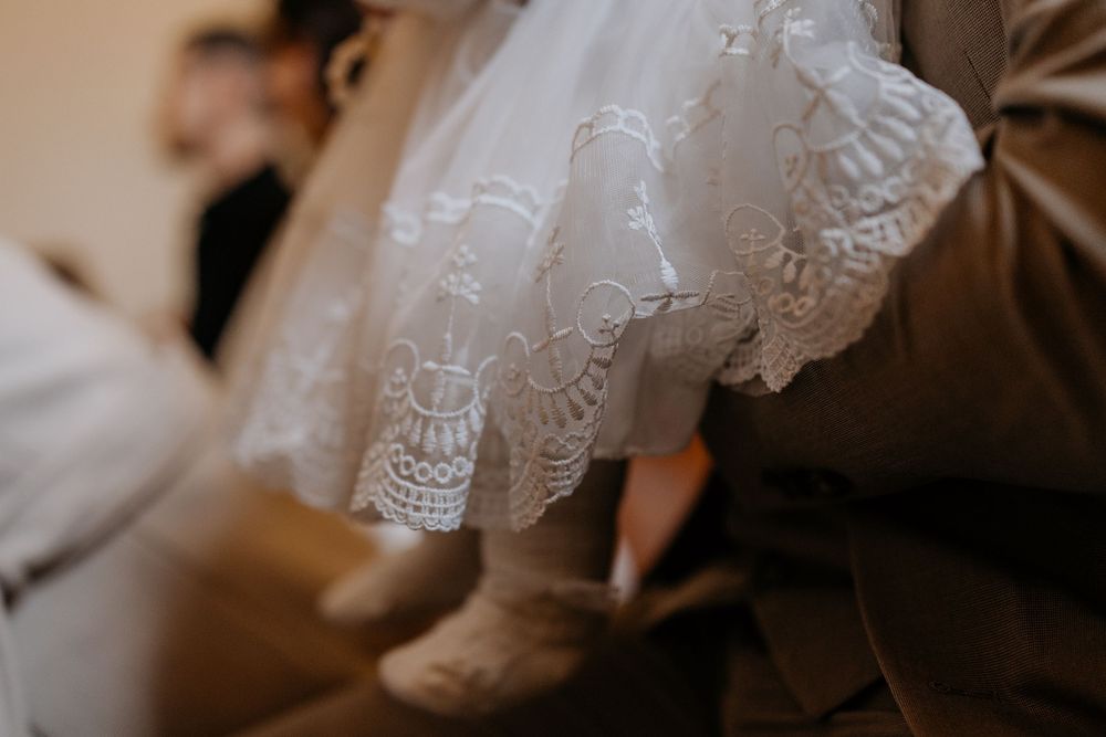 Close-up of a baby in a white lace christening gown with tiny booties resting on an adults lap.