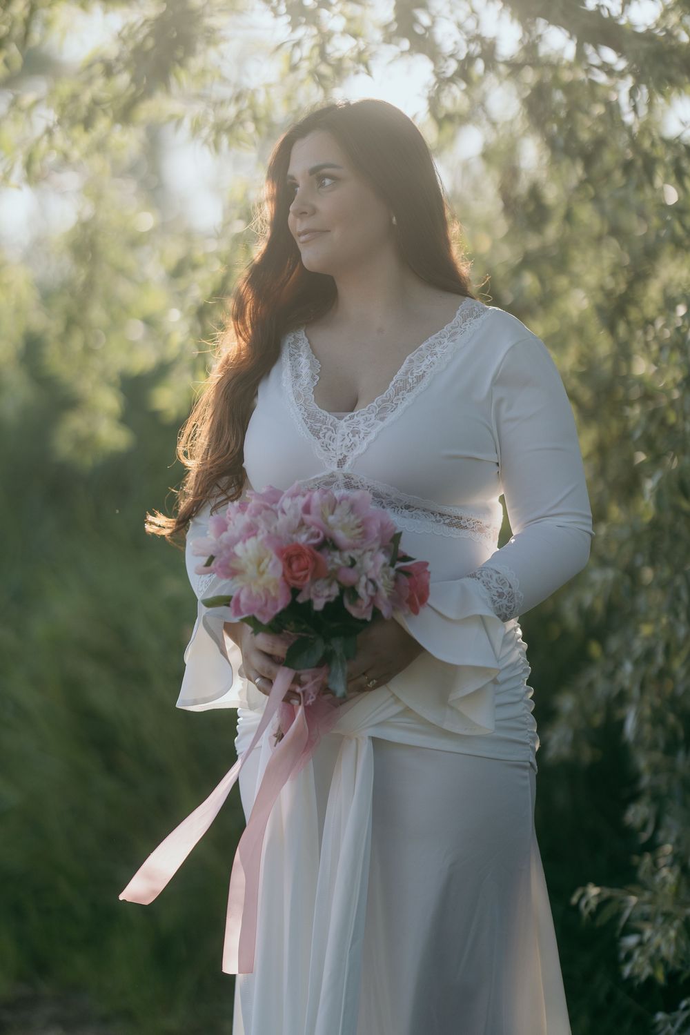 Pregnant woman in white lace dress holding pink flower bouquet outdoors at sunset.