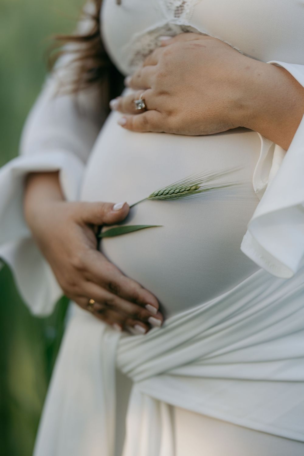 Pregnant woman in white dress cradling baby bump, holding a green wheat sprig outdoors.