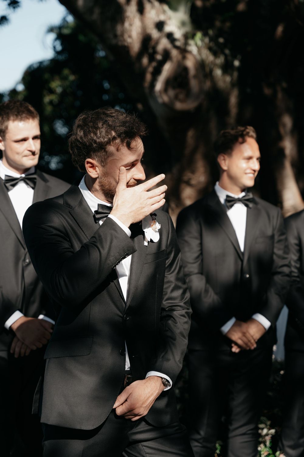Emotional groom covers his mouth while standing with smiling groomsmen in black tuxedos outdoors.