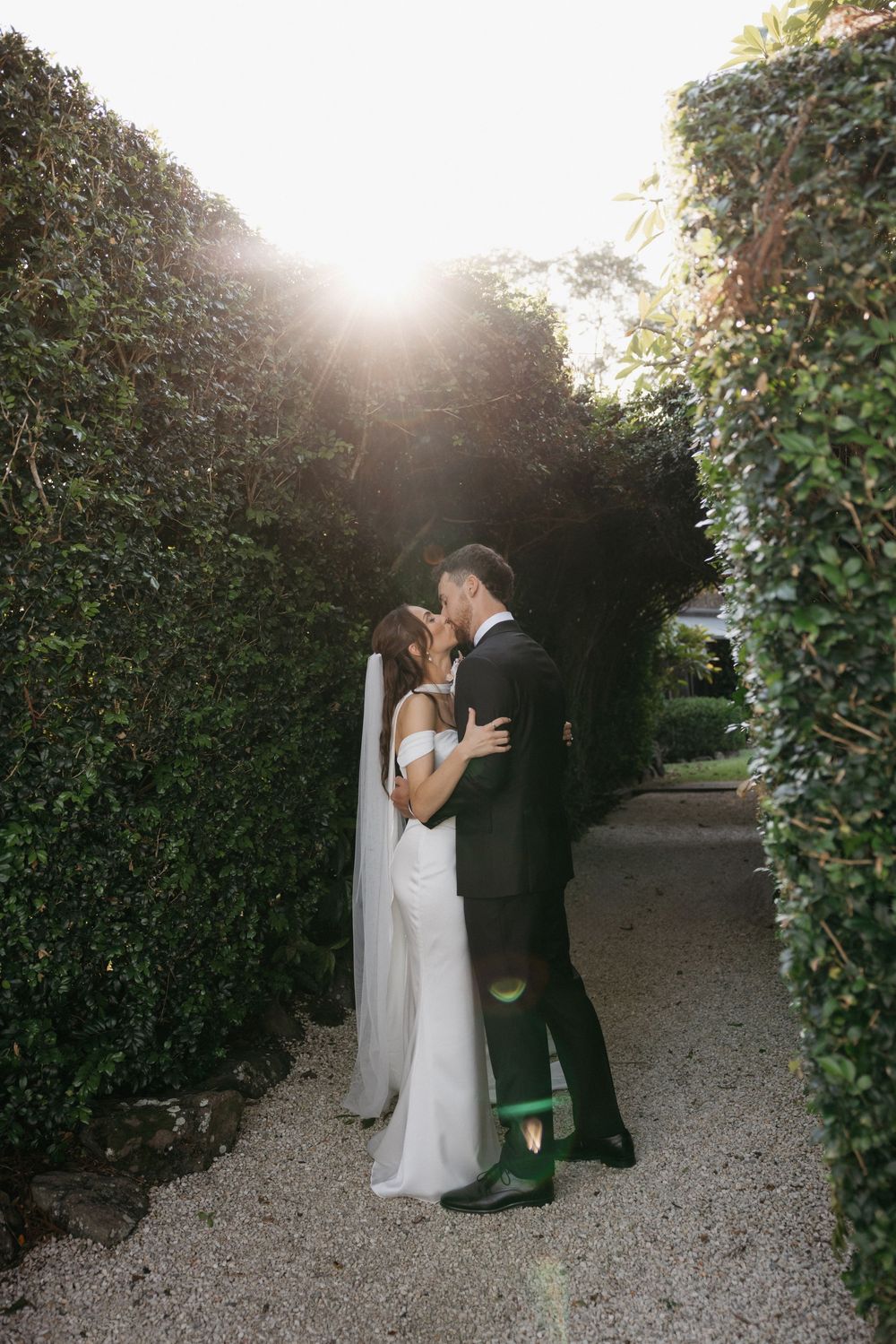 Bride and groom sharing a kiss in a sunlit garden hedge pathway on their wedding day.