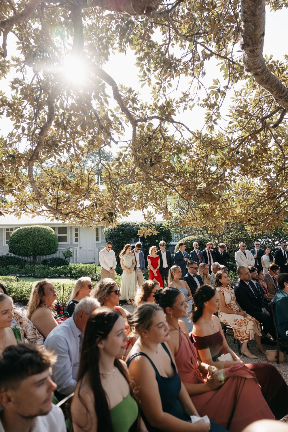 Outdoor wedding ceremony with seated guests under sunlit trees as the wedding party stands at the altar.