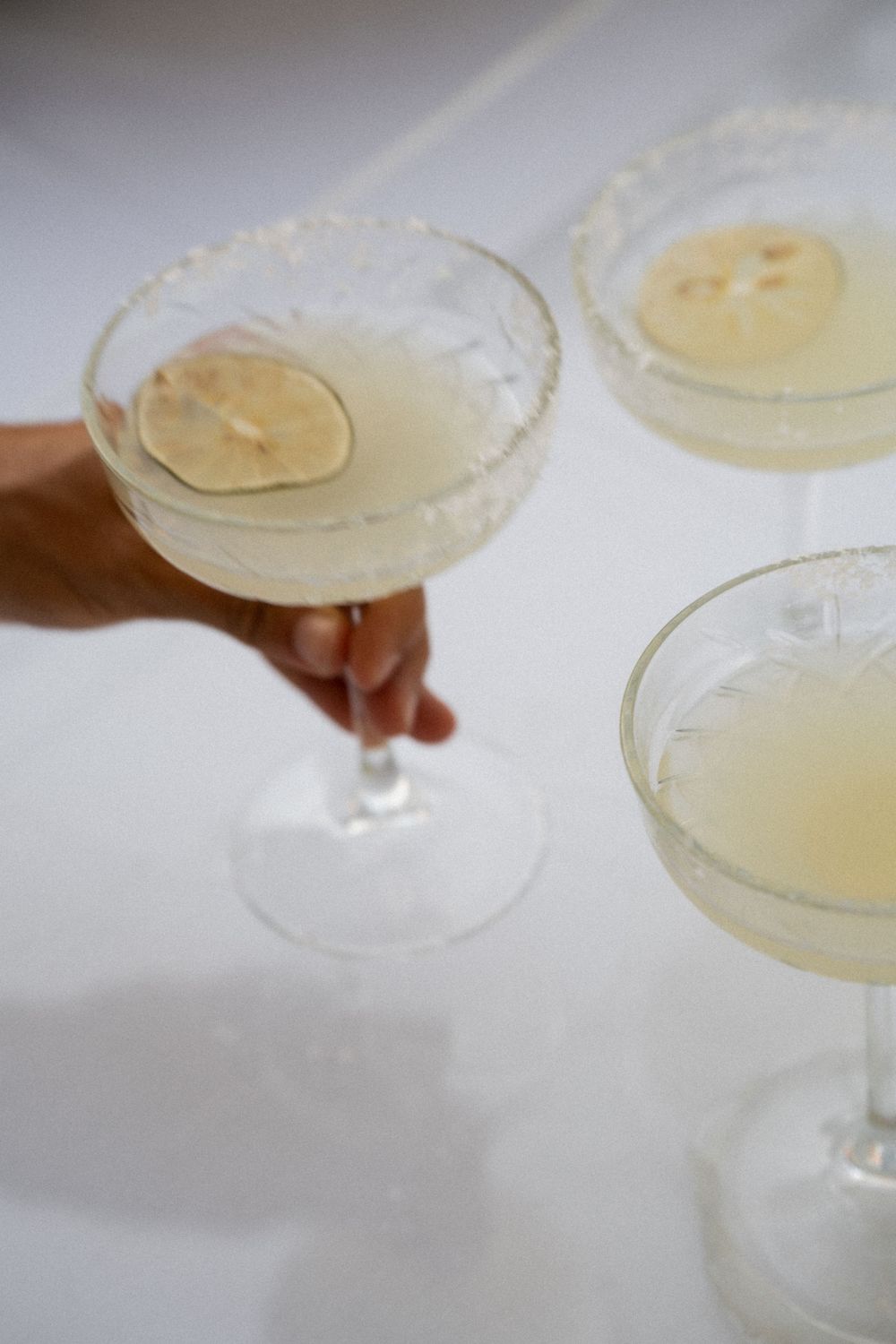 Hand reaching for a frosted coupe glass filled with pale yellow cocktail garnished with a dried lime slice.
