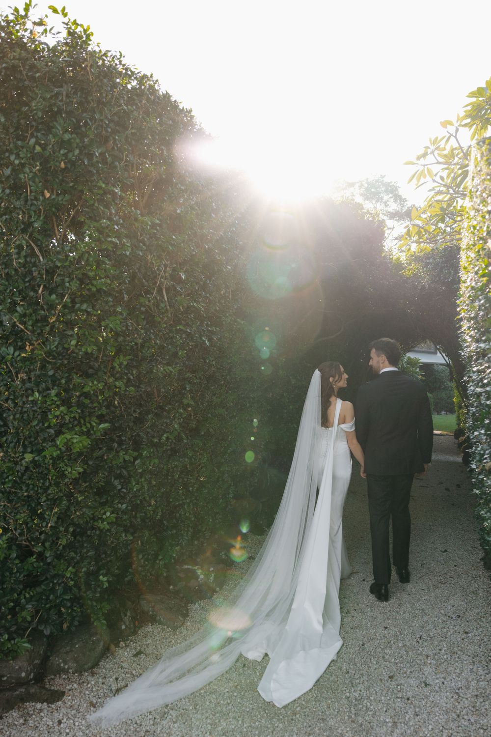 Bride and groom walking hand in hand through a sunlit garden hedge pathway in elegant wedding attire.