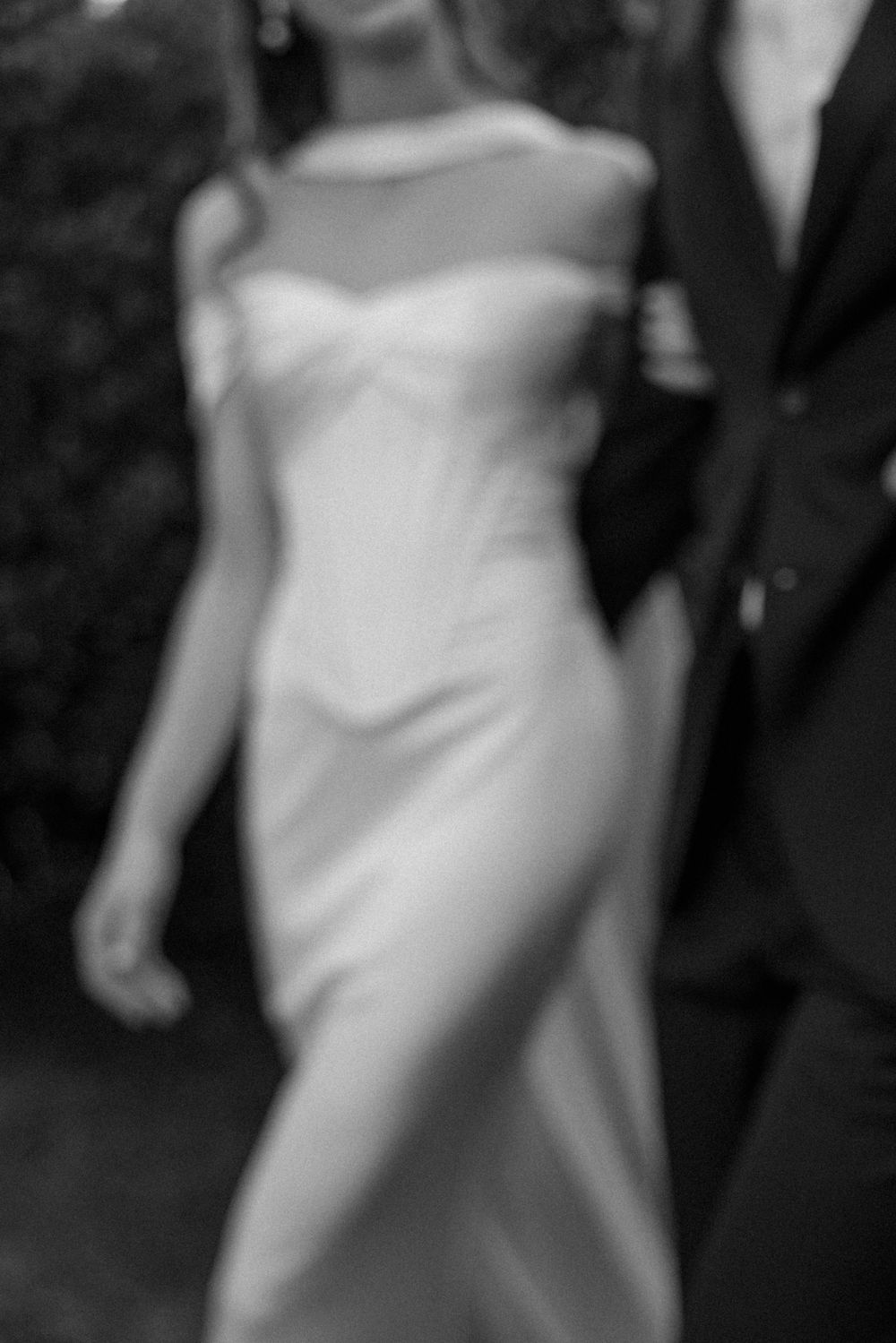 Blurred black and white photo of a bride in a sleek satin gown walking beside a groom in a dark suit.