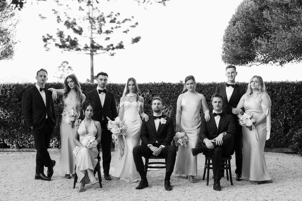 Wedding party posing in black and white portrait with bride, groom, groomsmen, and bridesmaids outdoors.