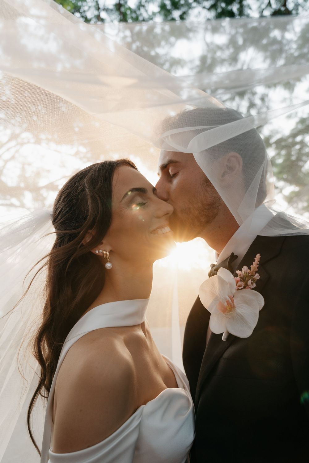 Bride and groom sharing a romantic kiss under a flowing veil with golden sunset backlight at outdoor wedding.