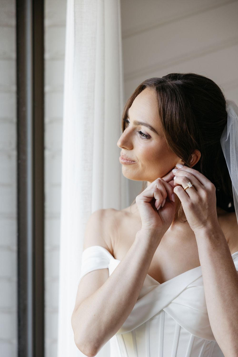 Elegant bride putting on earrings in white off-shoulder gown with veil by window.