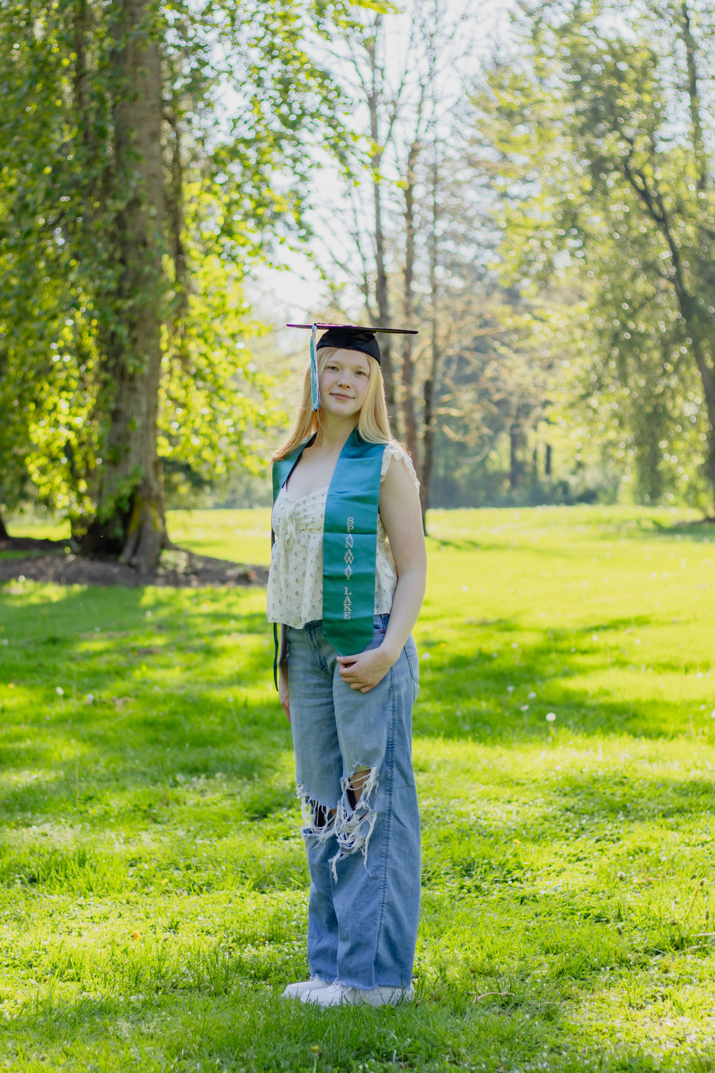 Smiling blonde senior in graduation cap, spanaway lake high school sash, and ripped jeans standing on lush grass taken by roy photographer