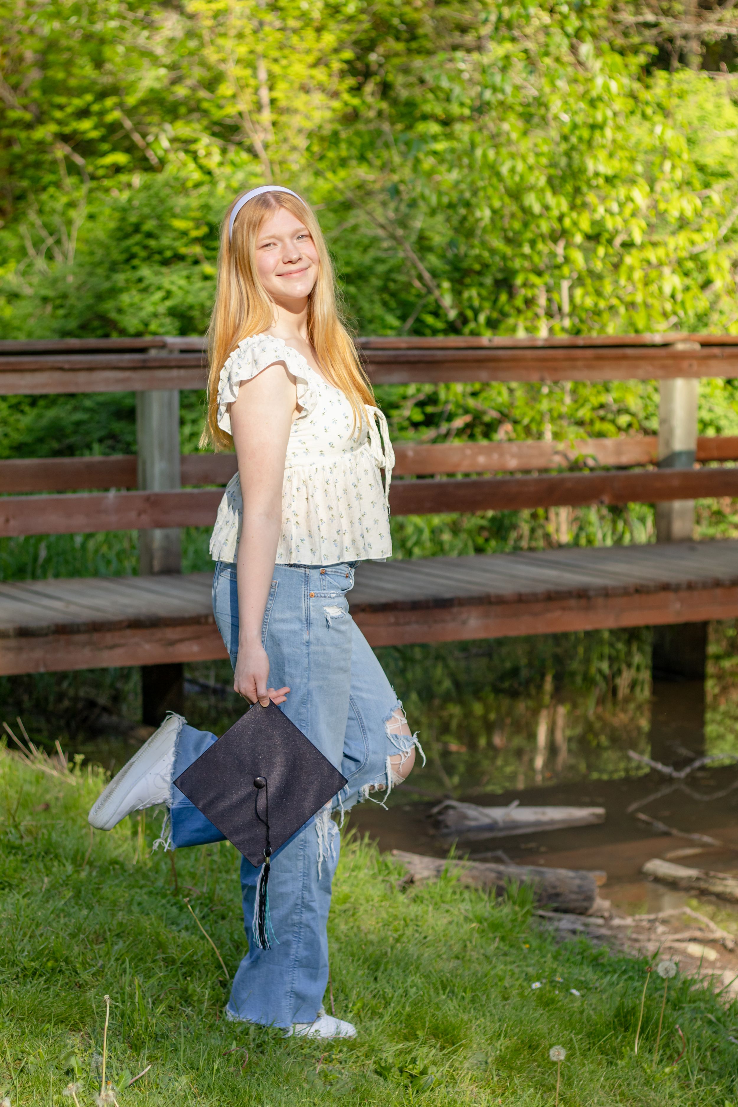 blonde teen in floral top holding a graduation cap near a wooden bridge at eatonville washington park taken by pierce county photographer