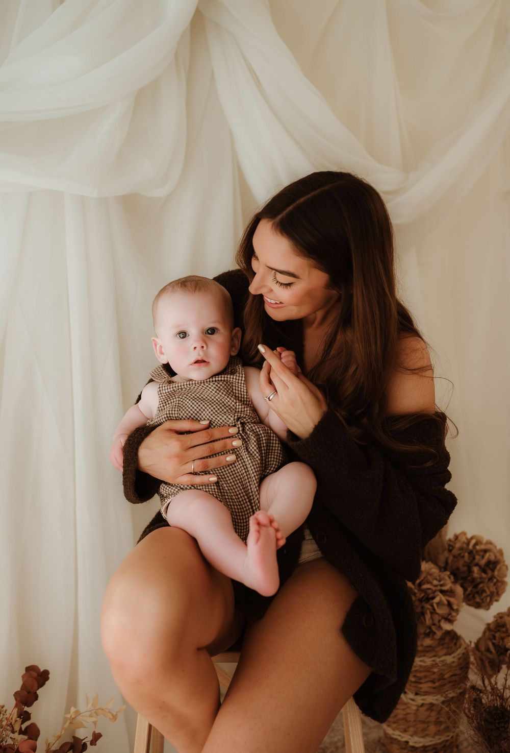 A young mother sits holding her baby in a soft, neutral-toned studio, both dressed in warm, earthy tones.