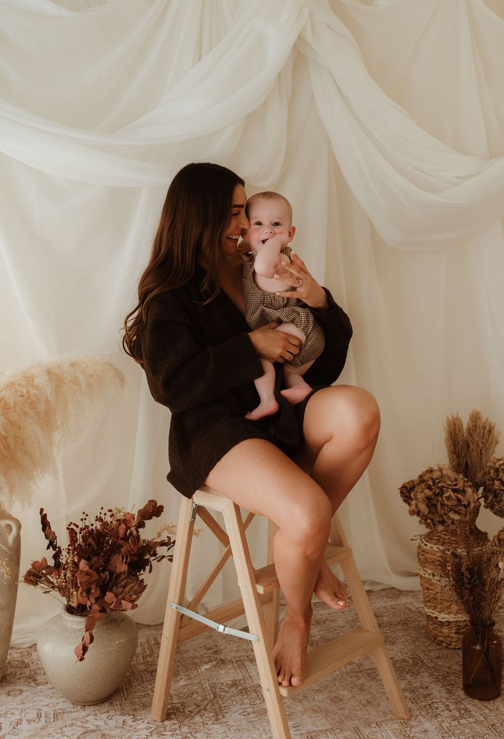 A woman sits on a wooden stool holding a baby, surrounded by dried florals and white draped fabric backdrop.