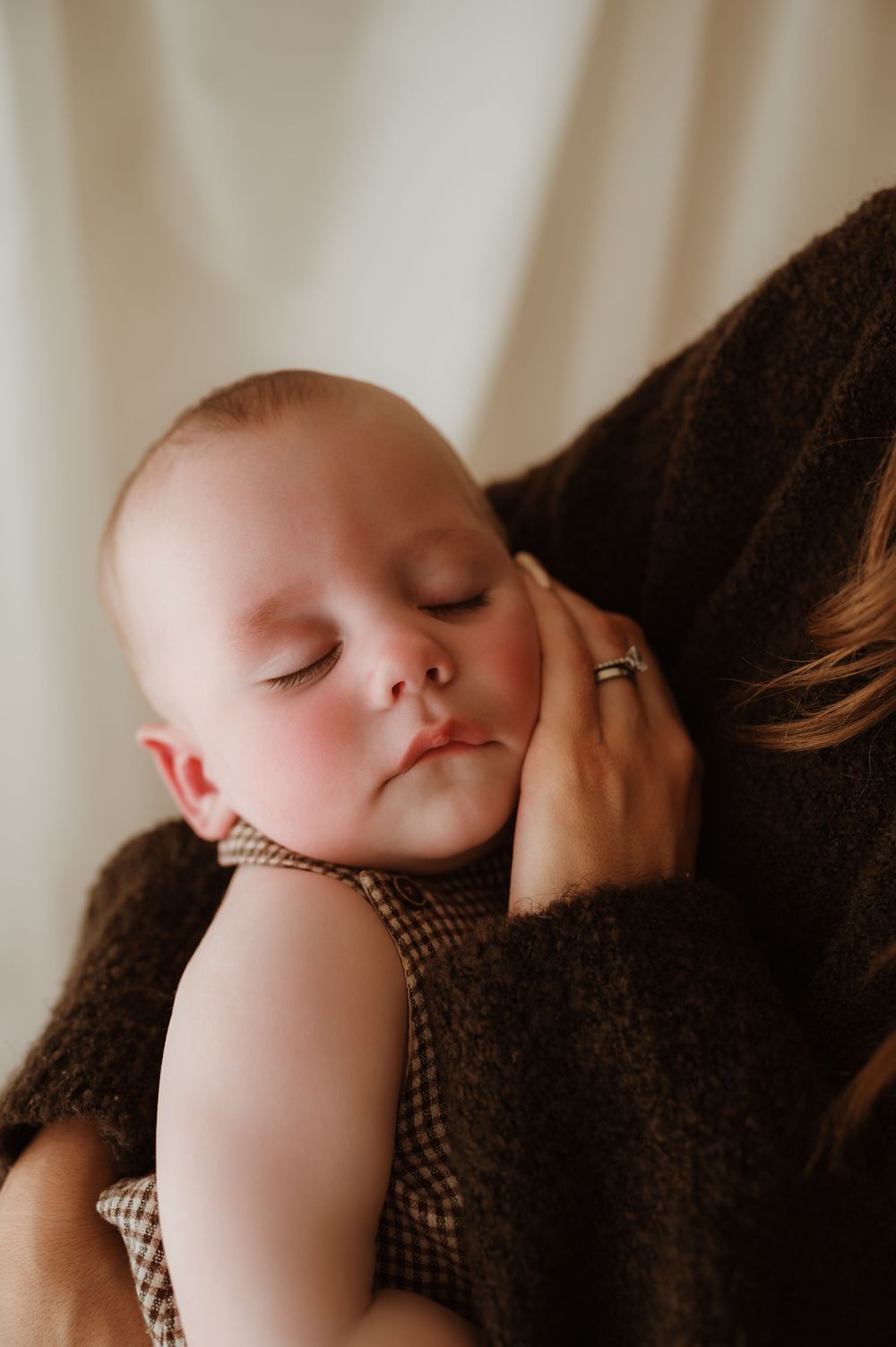 A sleeping newborn baby rests peacefully on a mother's shoulder, wearing a plaid outfit, as she holds them gently.