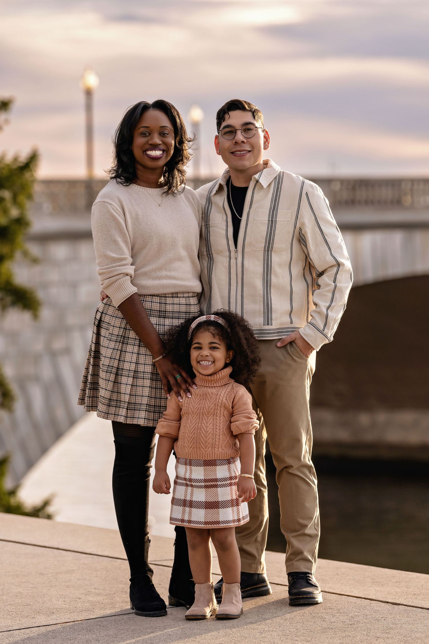 A joyful family of three poses outdoors near a bridge at sunset, with parents smiling behind their young daughter.