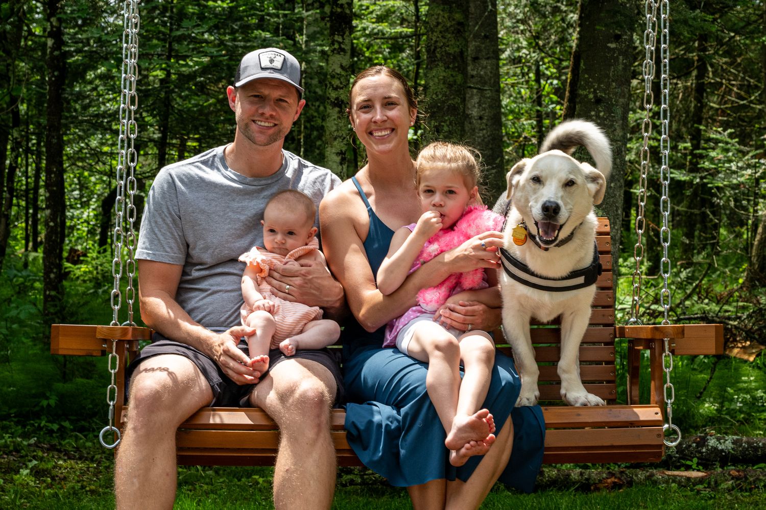 Family of two adults with baby and toddler sitting on wooden swing with white dog in green forest.