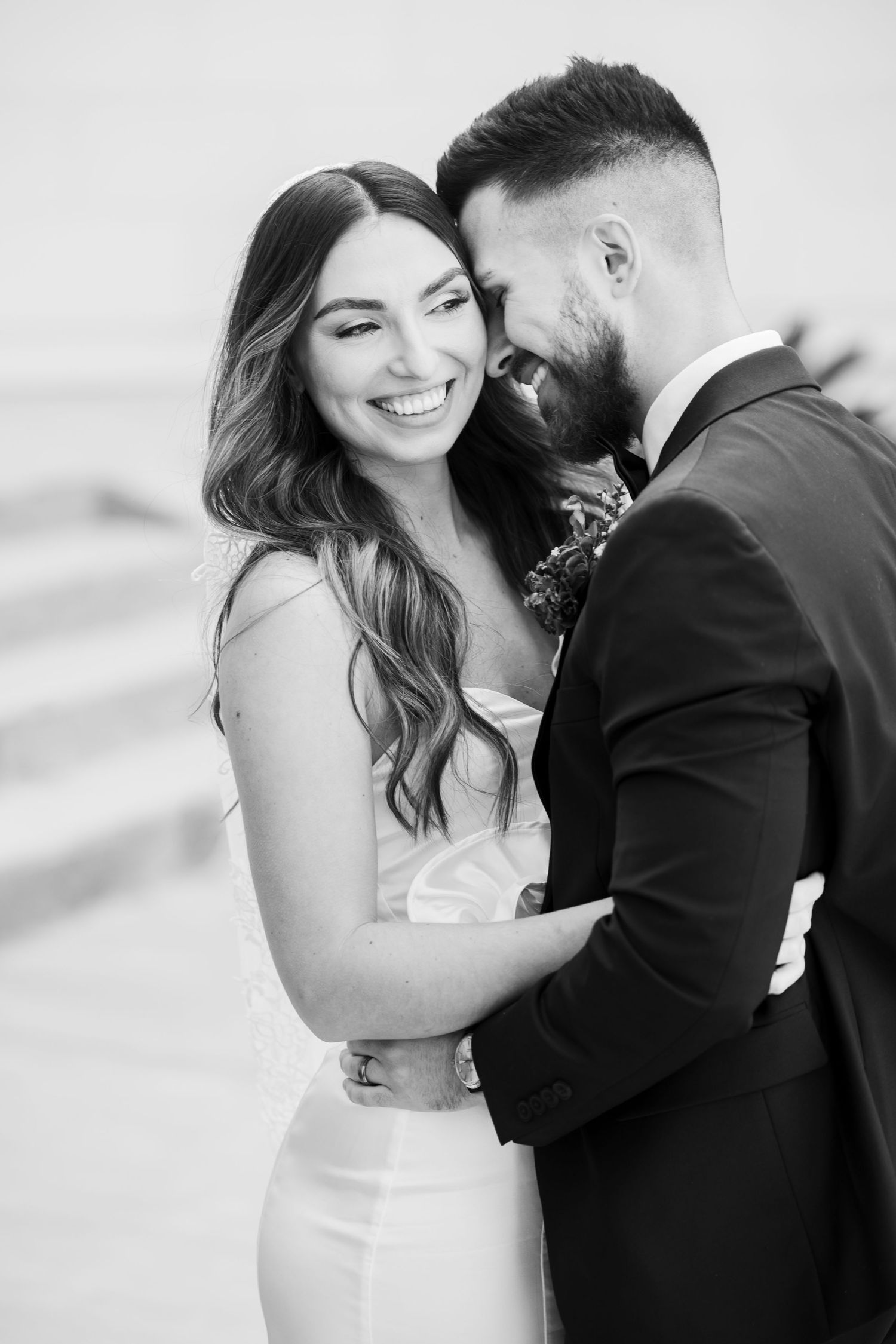 Black and white wedding photo of smiling bride in white gown embraced by bearded groom in dark suit.