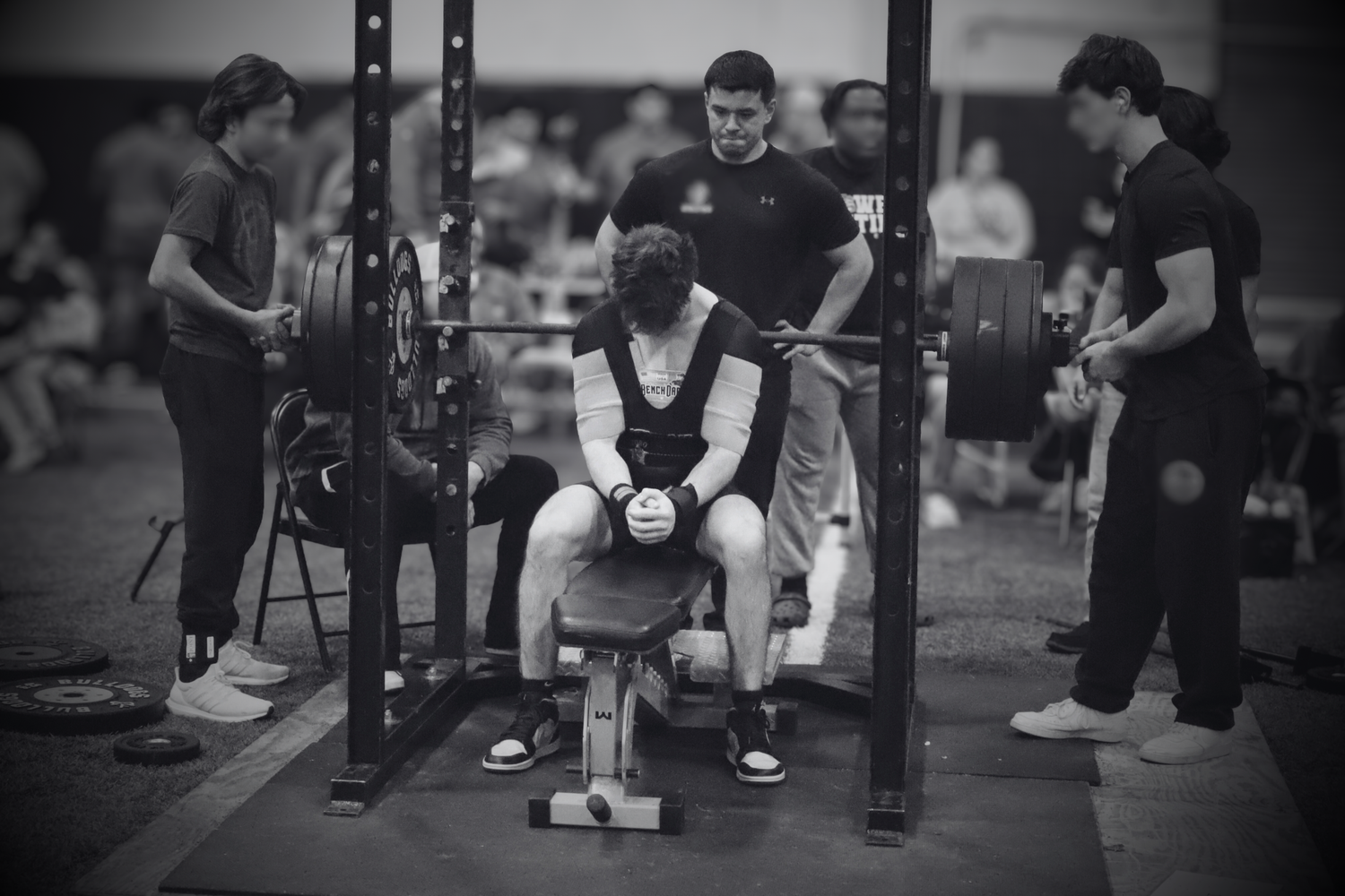 Black and white photo of athlete performing bench press with spotters at weightlifting competition.