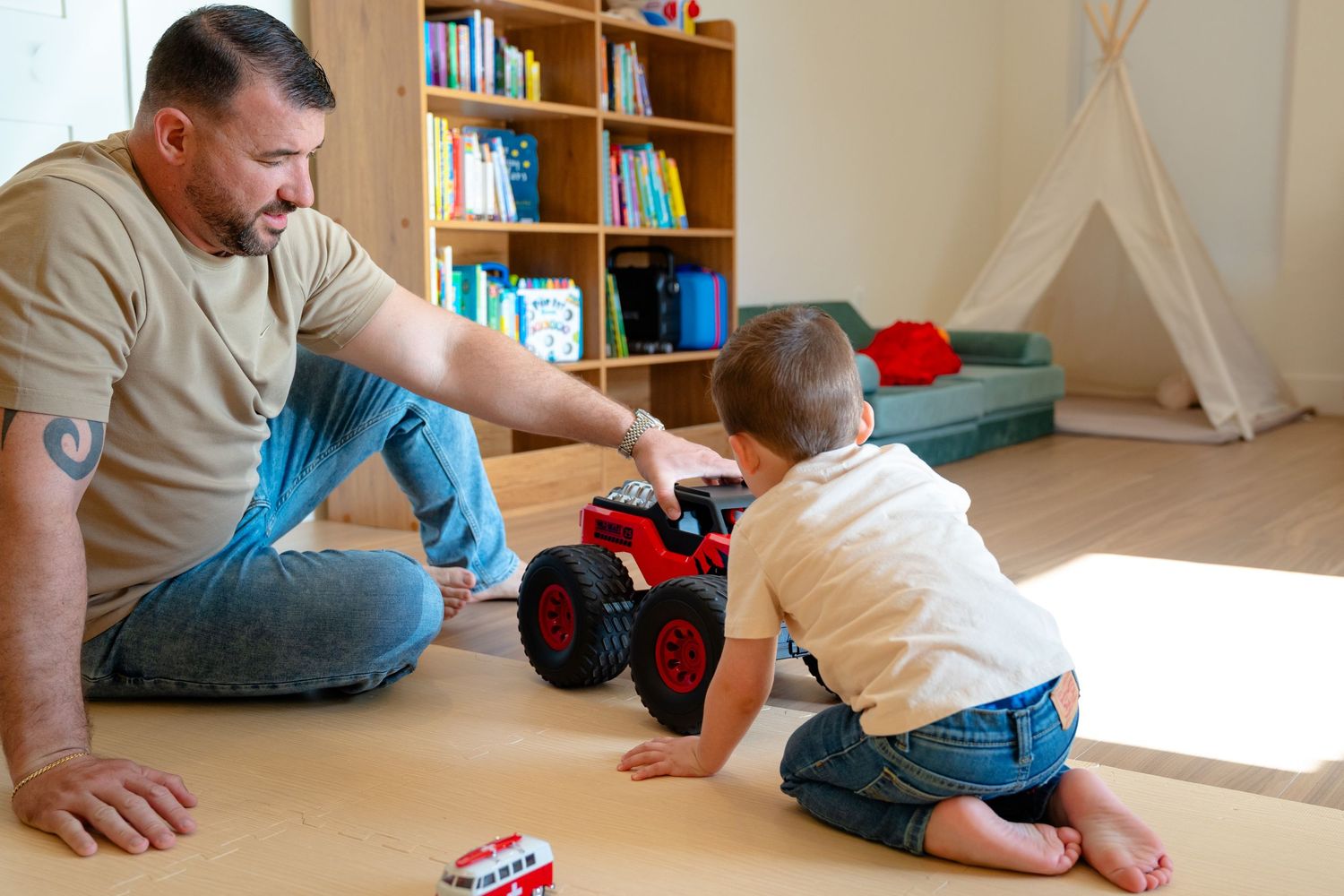 Father and toddler son playing with red toy monster truck on floor in colorful children's playroom.