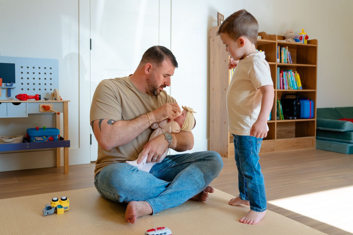 Father sits on floor playing with toy bear while young toddler boy stands nearby in bright children's playroom.