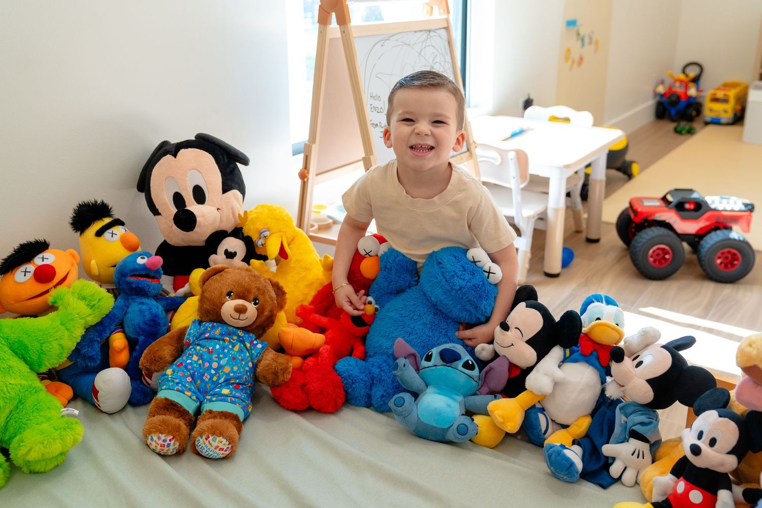 Smiling toddler surrounded by colorful stuffed animals and Disney plush toys on a white bed in a bright playroom.