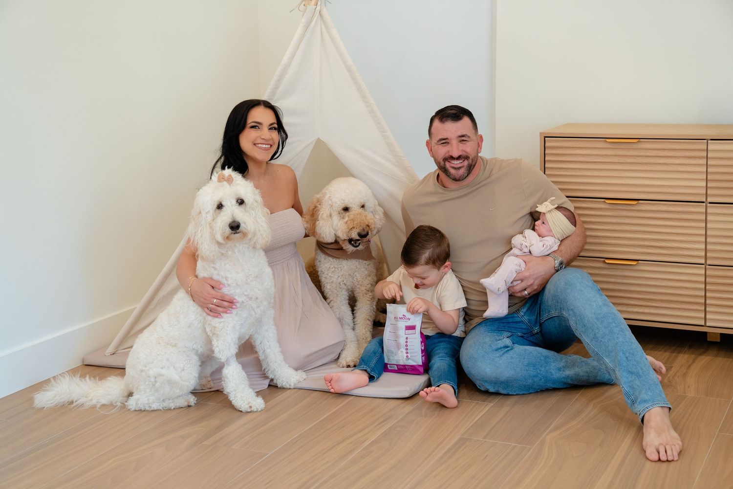 Happy family with two fluffy dogs, a toddler, and a newborn baby sitting together near a white teepee indoors.