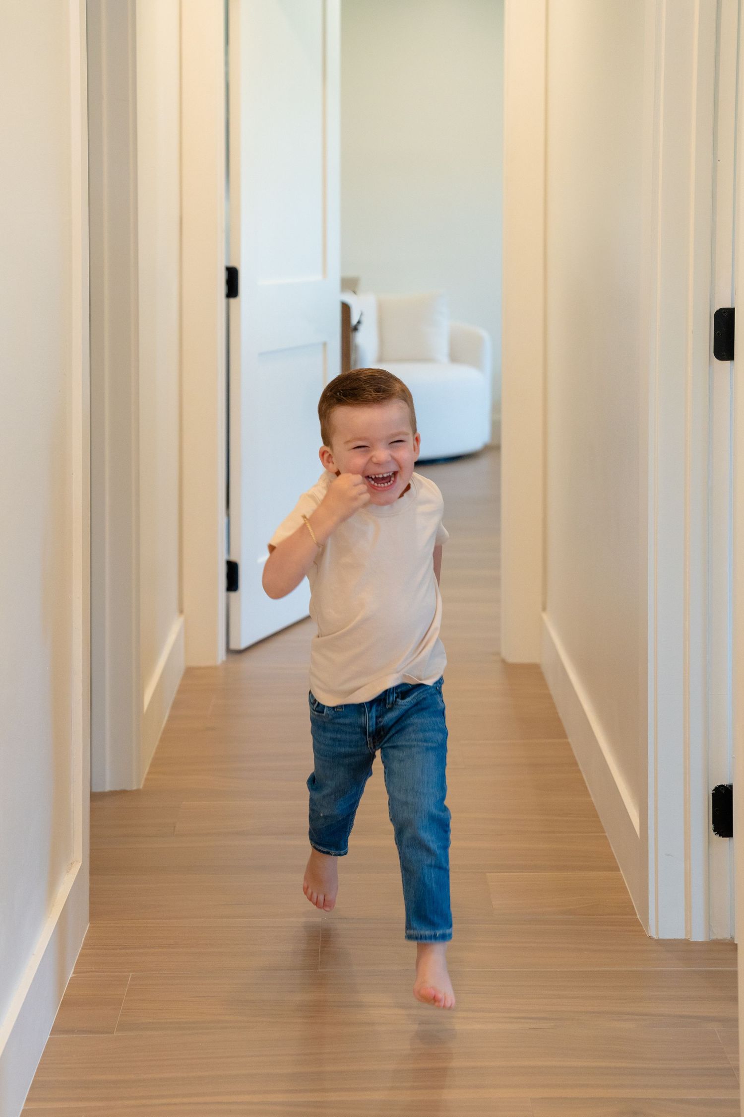 Happy toddler boy running barefoot down a bright home hallway, wearing jeans and a white t-shirt.
