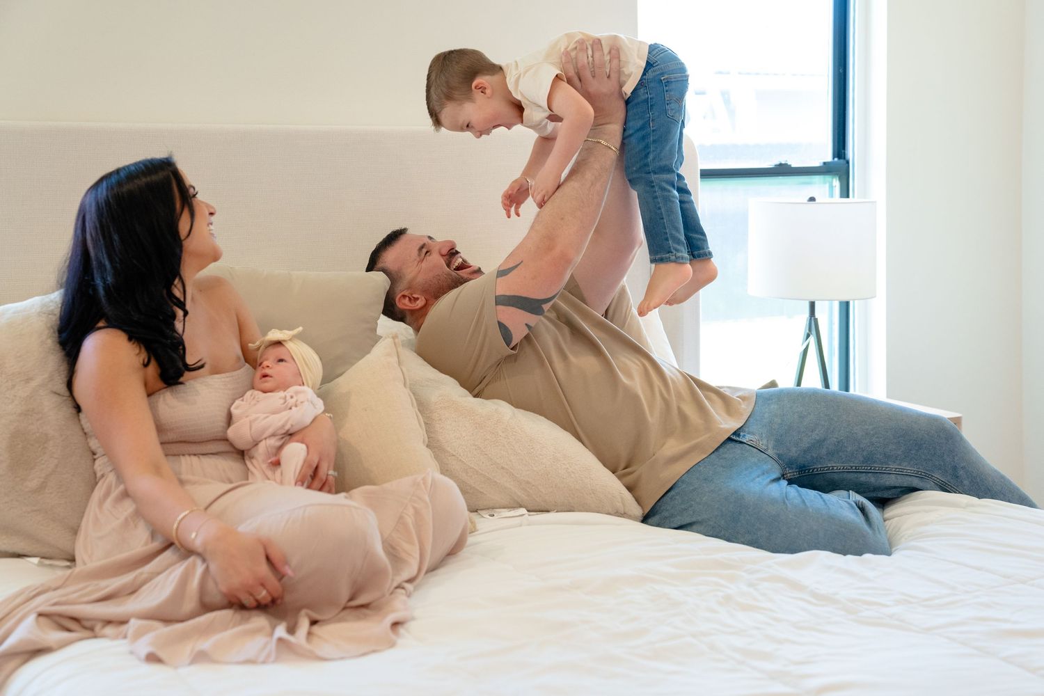 Family relaxing on a white bed, mother holding newborn baby while father plays with toddler child in bright bedroom.