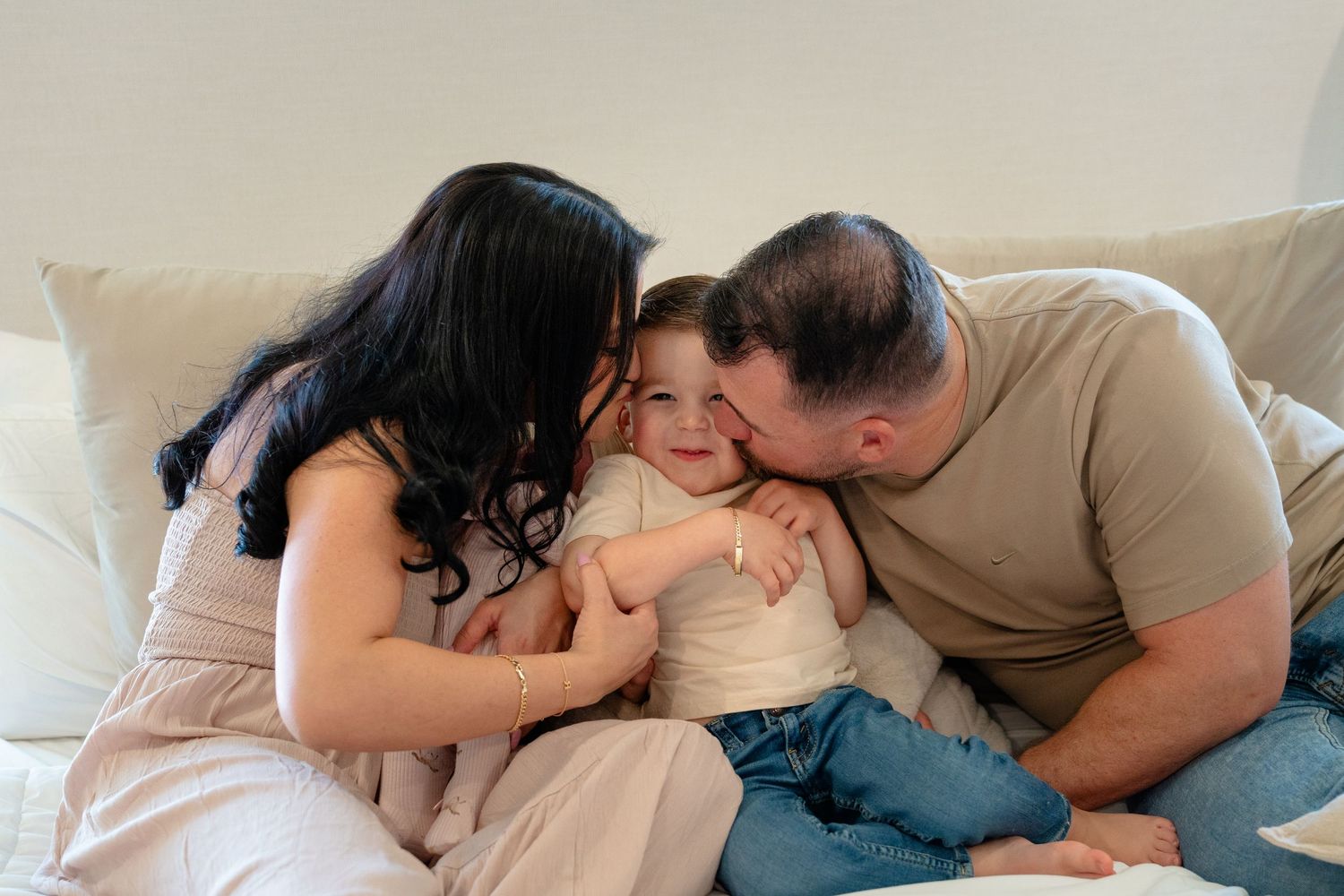 A happy family with two parents kissing and cuddling their smiling toddler on a cozy white couch.