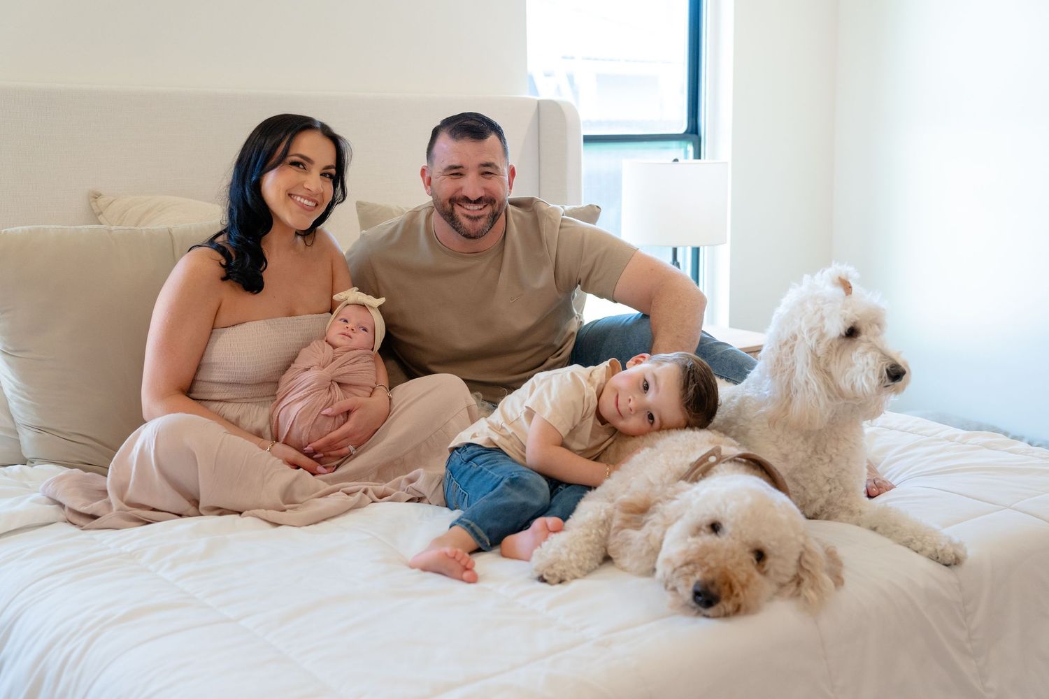 Happy family with newborn, toddler, and fluffy white dog relaxing together on a white bed at home.