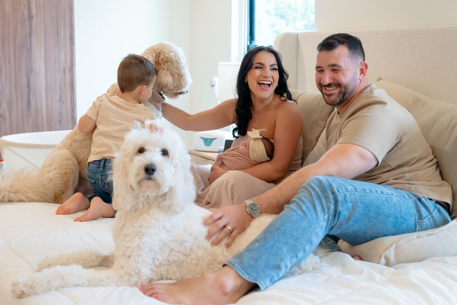Happy family with two fluffy white dogs relaxing on a bed, smiling and enjoying a cozy moment together at home.