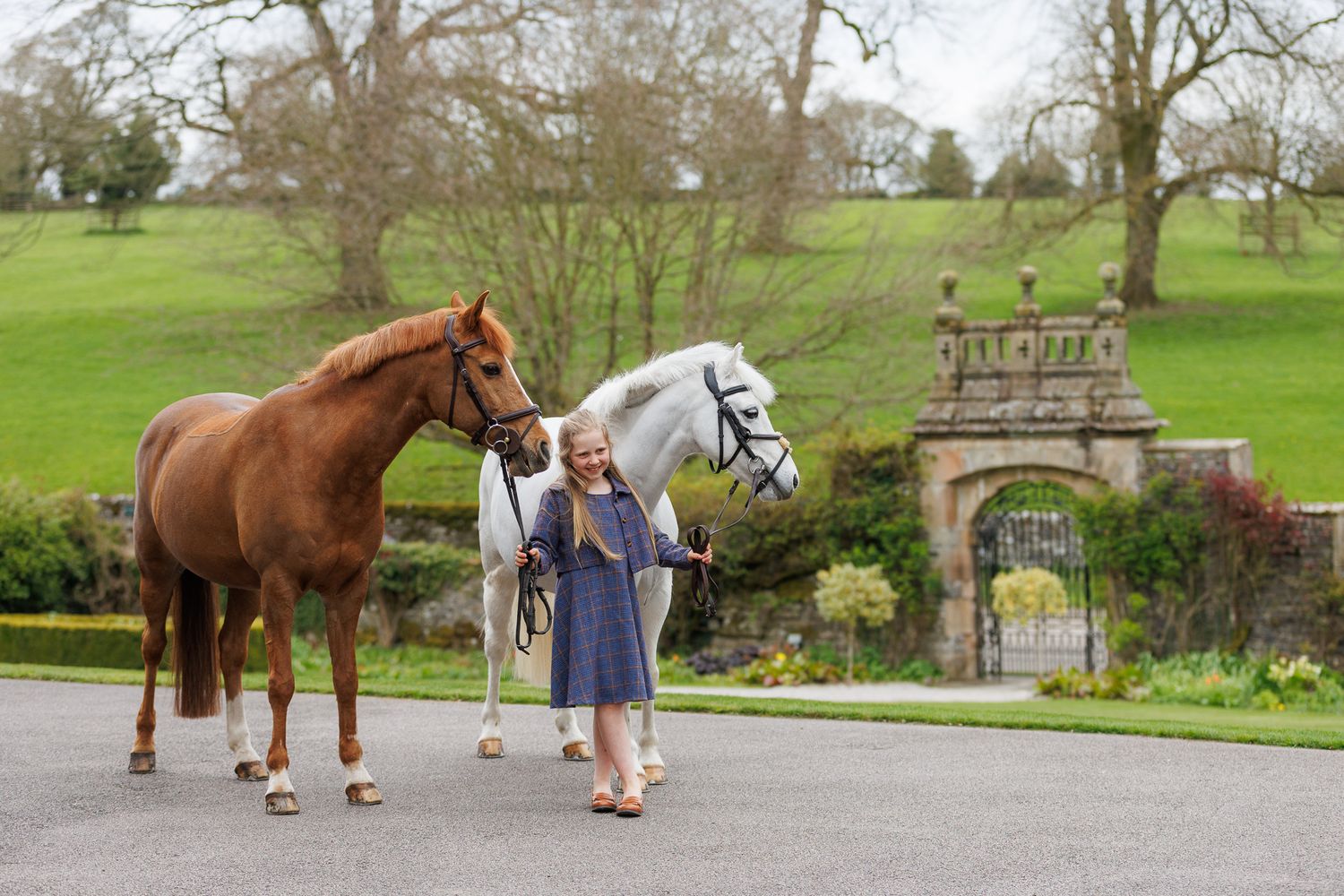 Young girl standing between a chestnut and grey horse near ornate stone gates in a lush green estate.