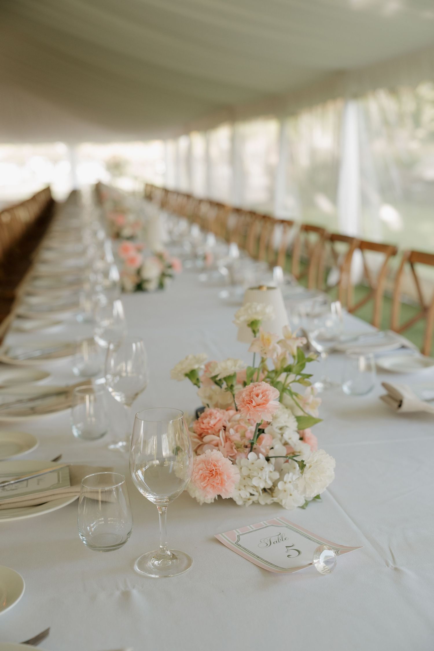 Elegant wedding reception table with blush pink floral centerpieces, white linens, and crystal glassware under a tent.