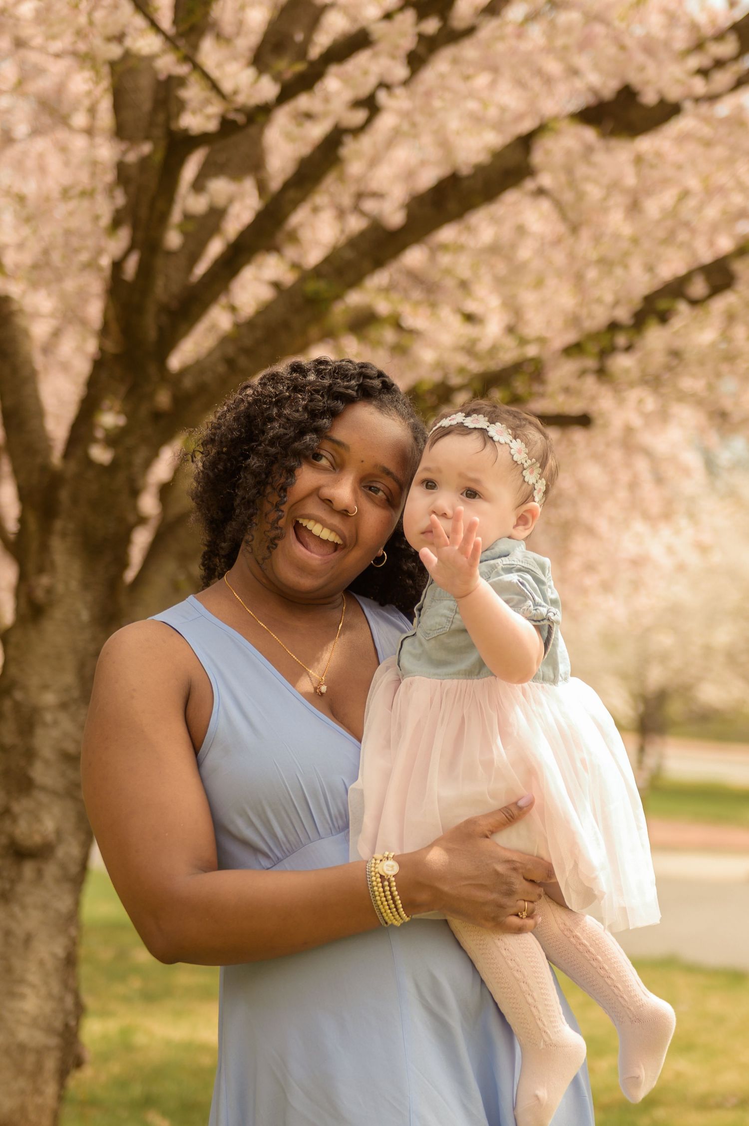 A joyful woman in a blue dress holds a baby girl in a pink tutu under blooming cherry blossom trees.