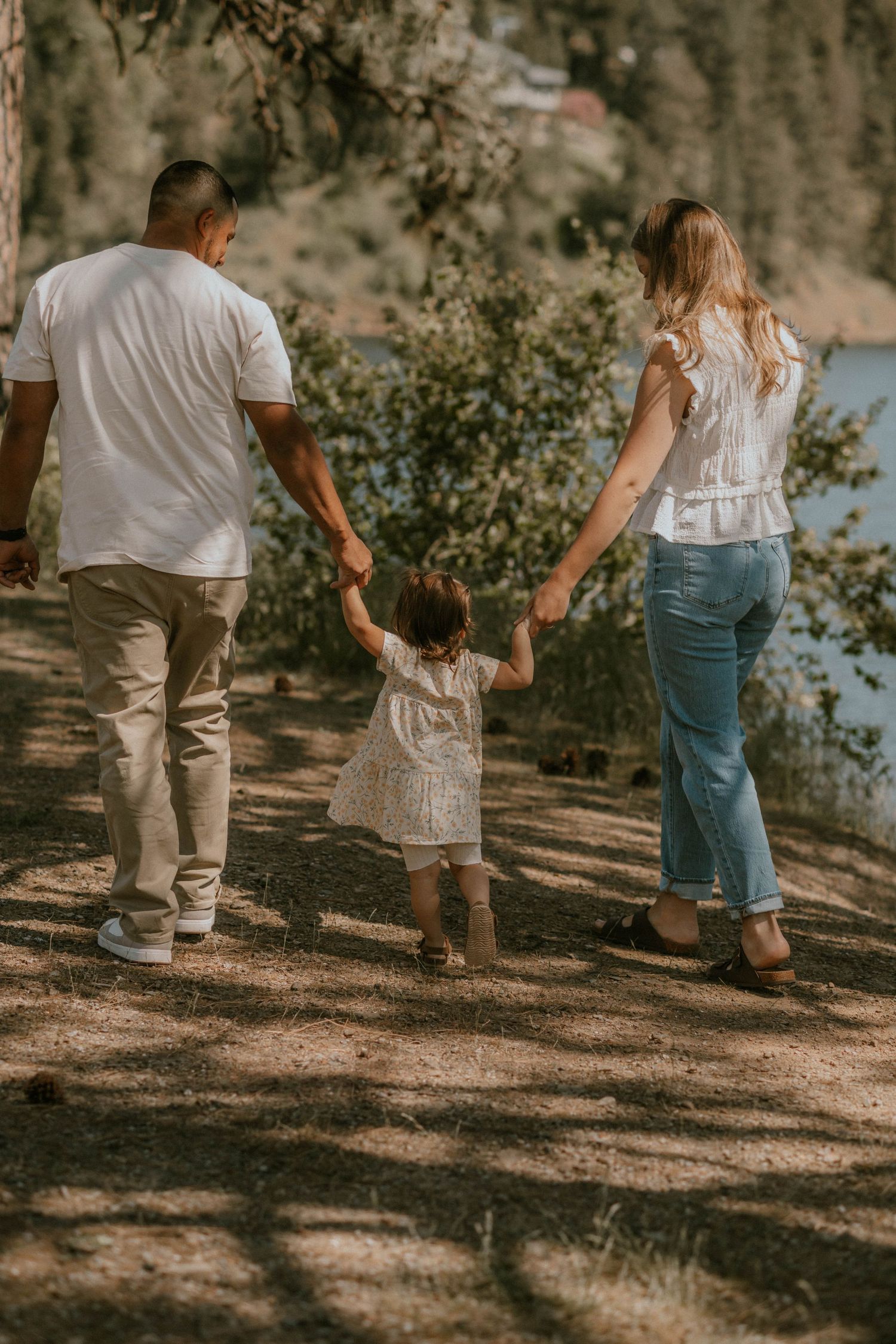 Family of three walking hand-in-hand on a sunny forest trail, parents guiding toddler in white dress.