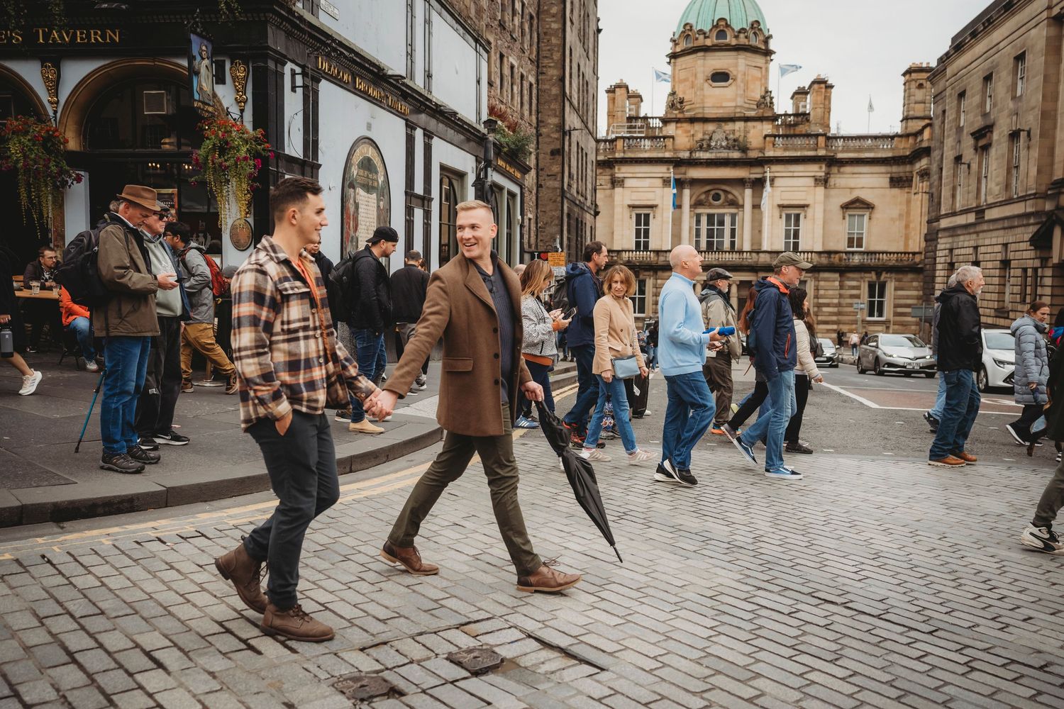 Two men hold hands and walk together on a cobblestone street in Edinburgh, Scotland, with historic buildings in the background.