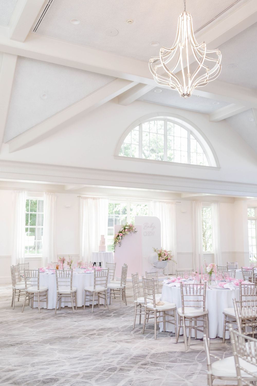 Elegant white ballroom with pink floral decor, chiavari chairs, round tables, and a modern chandelier for a wedding reception.