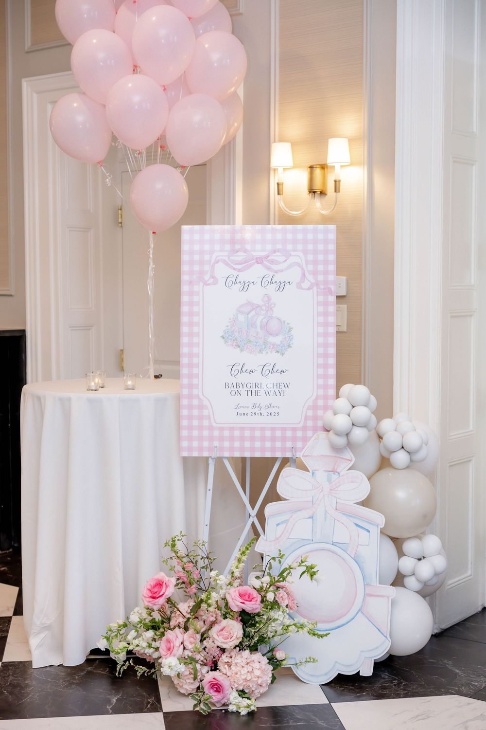 Pink baby shower display with gingham welcome sign, floral arrangement, balloon arch, and decorative cutouts in elegant venue.