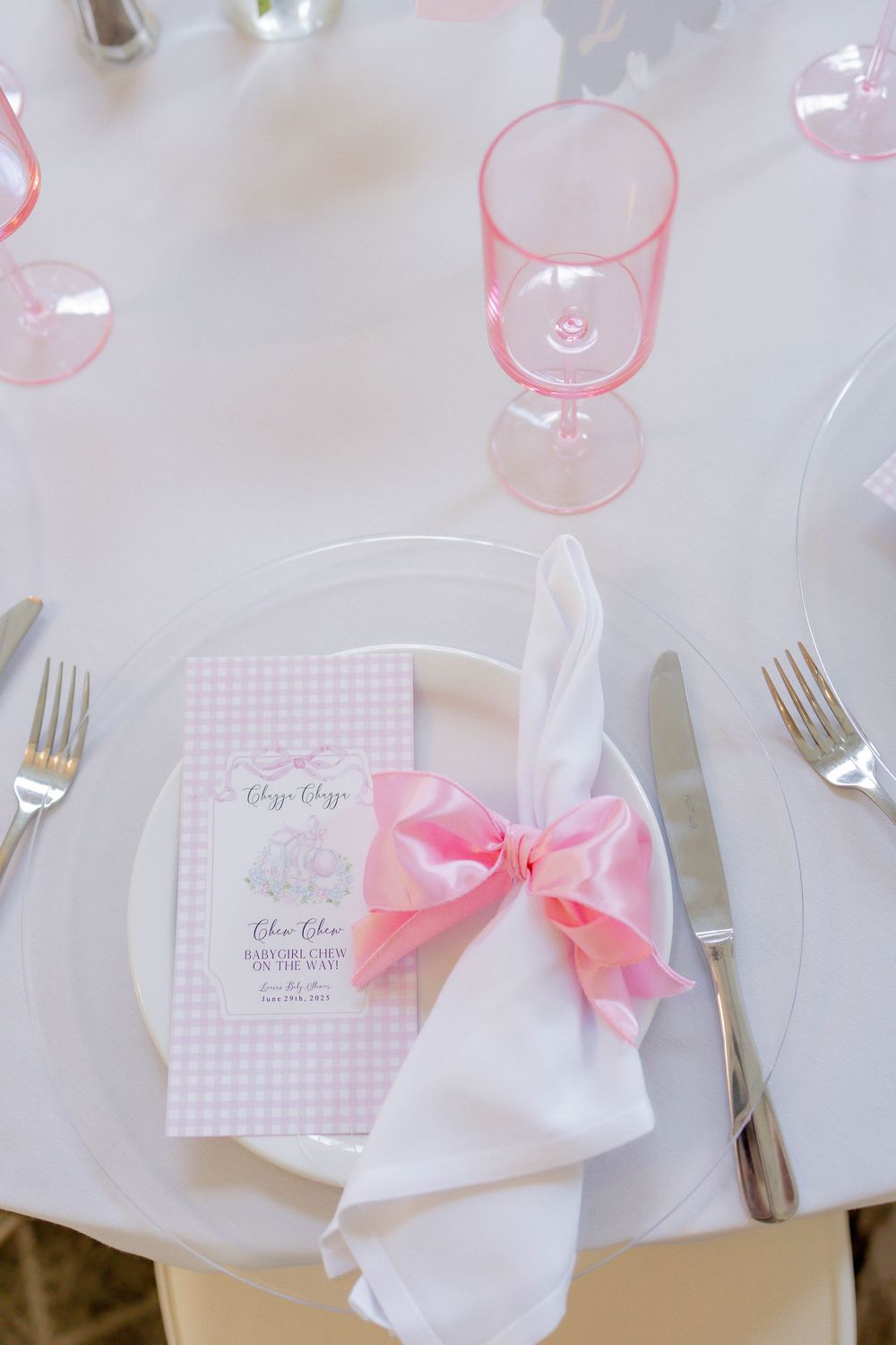 Elegant baby shower place setting with pink gingham menu card, white napkin tied with pink bow, and pink stemware.