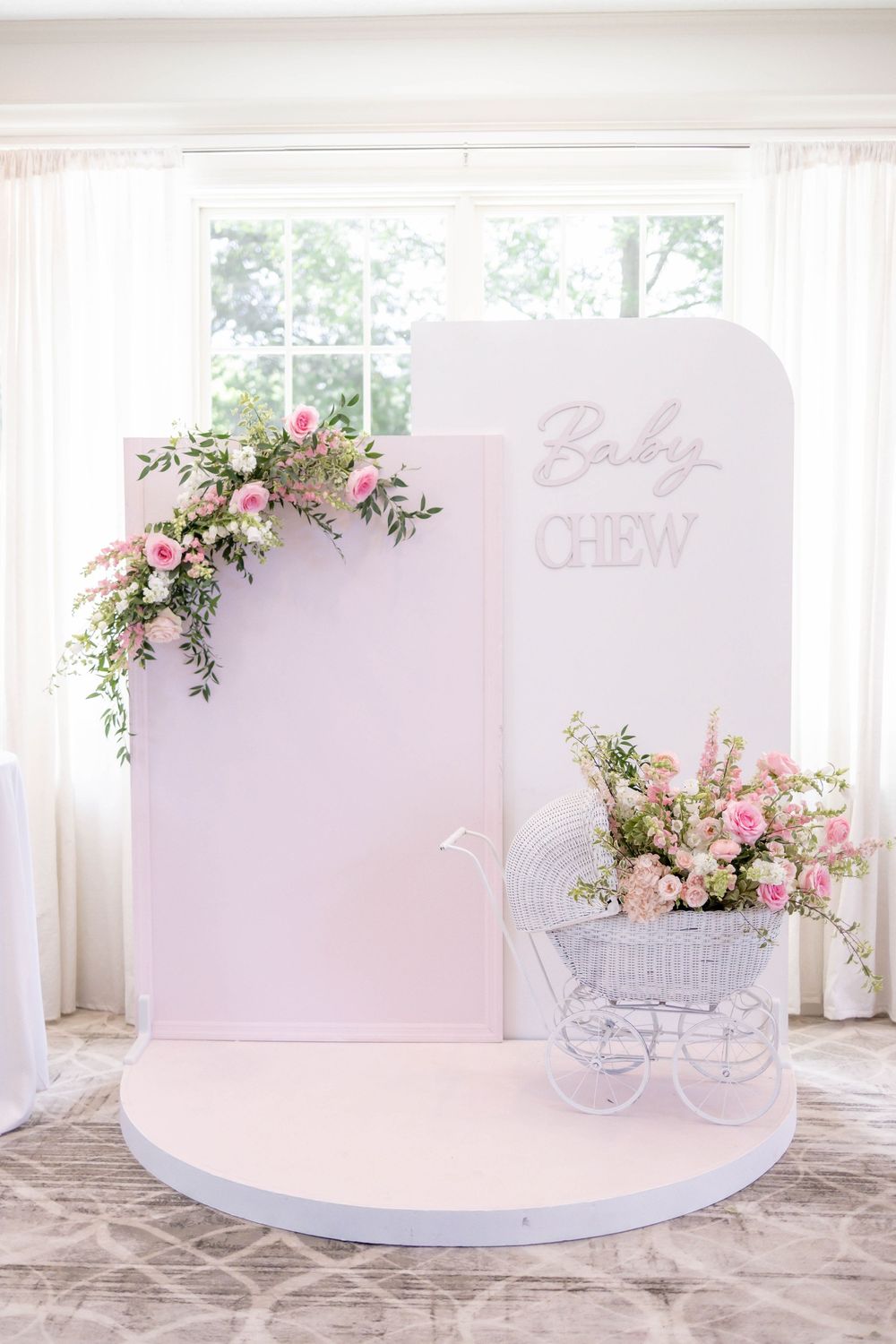 Baby shower backdrop with pink floral arch, white panels, and vintage white pram with pink flowers.