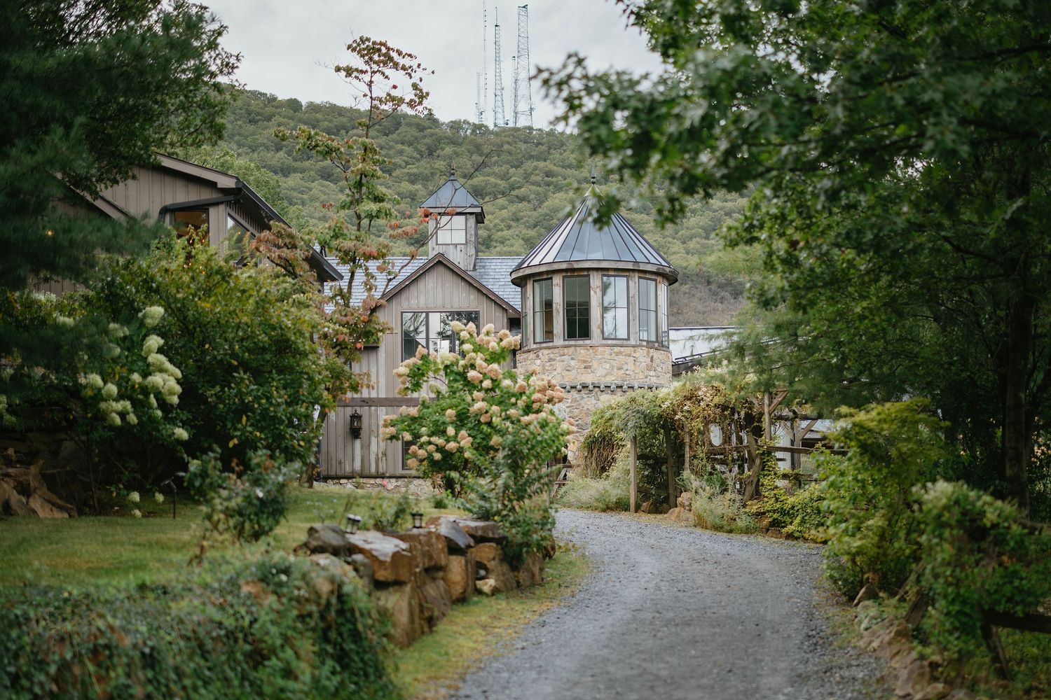 Charming stone cottage with turret surrounded by lush greenery and blooming flowers along a gravel path.