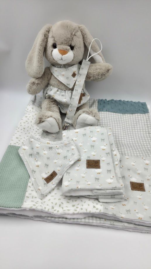 Soft plush gray bunny rabbit stuffed animal toy with floppy ears and floral bib, seated on white blanket.