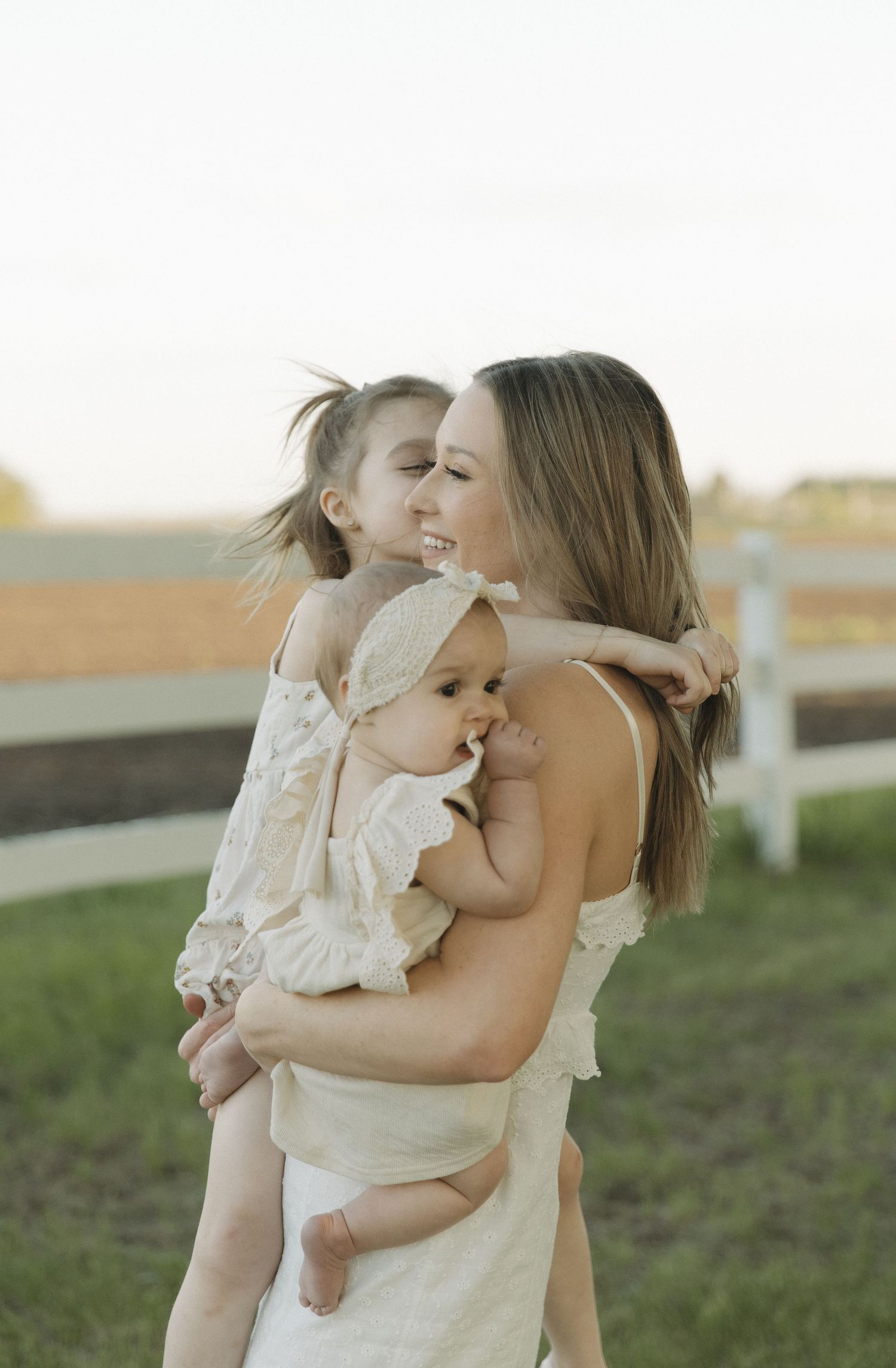 A mother in a white dress smiles while holding her baby and hugging her young daughter outdoors at golden hour.